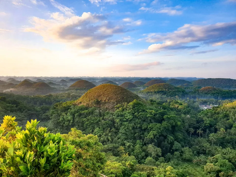 Rounded grassy hills rise above dense green trees beneath blue sky and pale clouds.