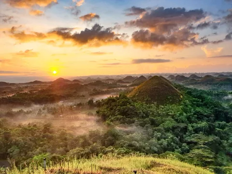 The rising sun lights clouds above the Chocolate Hills, with mist between green slopes and trees.