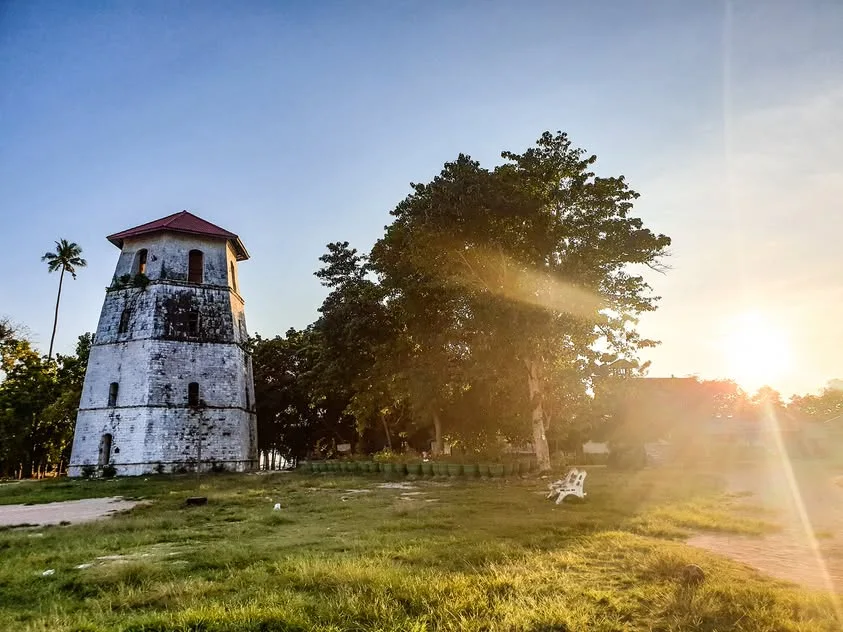 A tall white tower with dark weathering and a red roof stands beside trees and a grassy area in low sunlight.