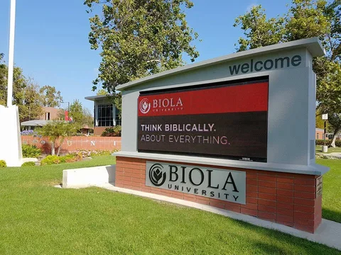 Biola University’s welcome sign stands on a green lawn beneath trees.