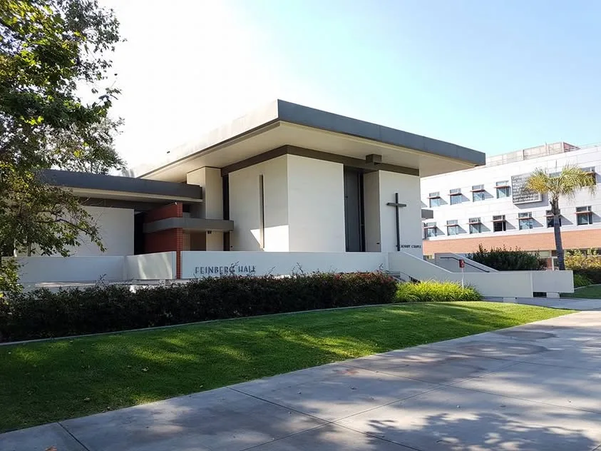 Calvary Chapel has white walls and a simple cross above a lawn, with Feinberg Hall lettering along the lower wall.