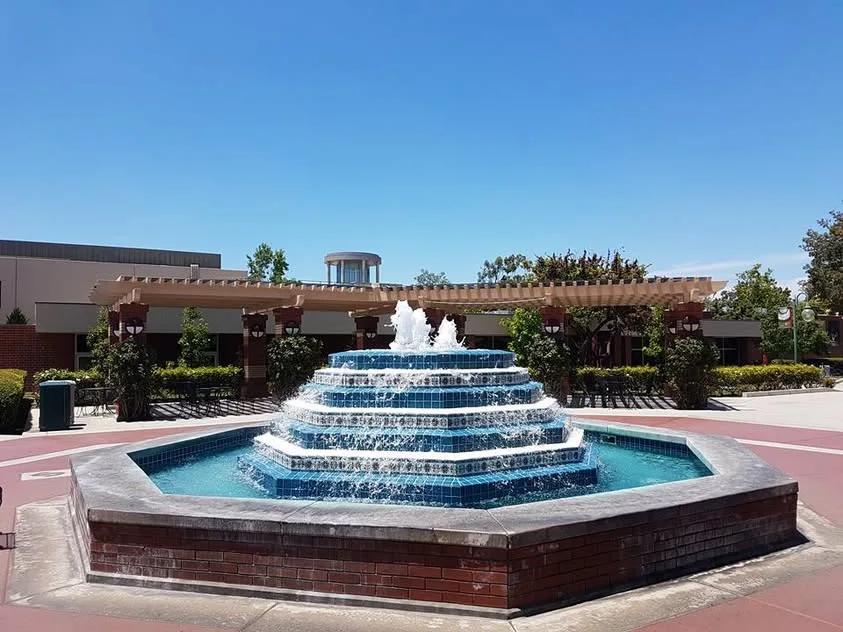 Water runs down a tiered blue-tiled fountain in a campus plaza, with a pergola behind it.