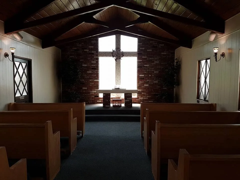 Empty wooden pews face an altar and a cross-shaped window beneath a dark timber ceiling.