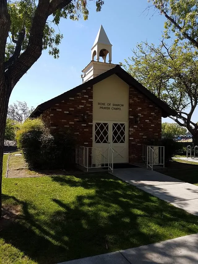 The small brick Rose of Sharon Prayer Chapel has a white bell tower and stands among leafy trees.