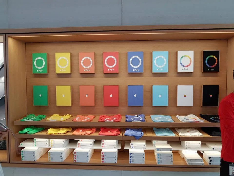 Colourful Apple Park notebooks and T-shirts arranged on wooden shelves; portions of visitors’ bodies are visible at the edges, with no faces.