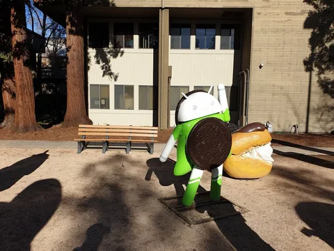 A green and white Android Oreo statue carries a giant cookie in a sunny courtyard beside a bench.