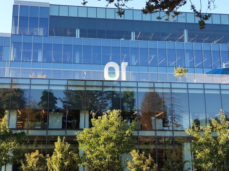 A white Android 10 display stands on a balcony behind a railing against the glass façade of an office building.
