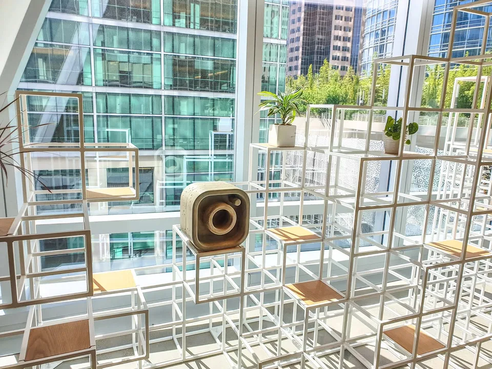 A camera-logo sculpture sits on open white shelving beside tall windows overlooking nearby buildings.
