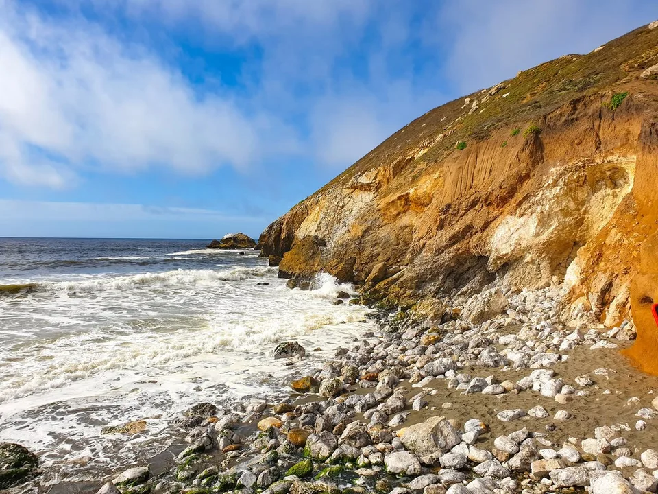 Foamy waves wash over a rocky shore beneath a steep golden-brown coastal bluff.