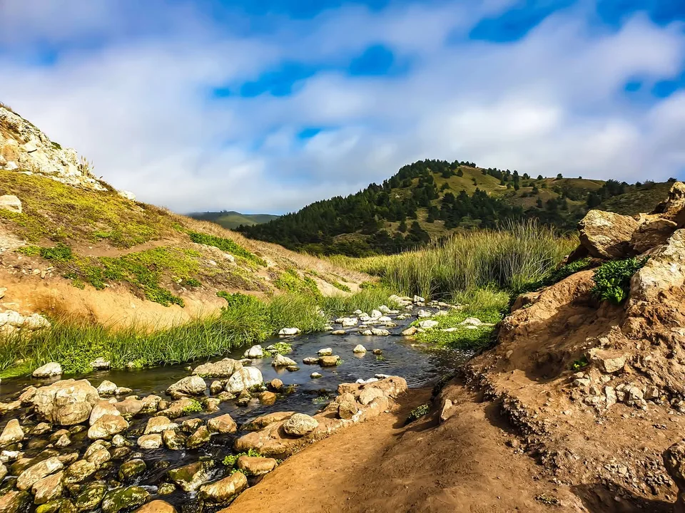 A shallow stream flows among pale rocks and grasses between coastal hills.