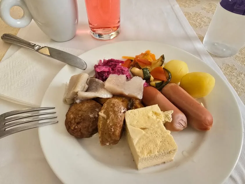 A breakfast plate holds fish, sausages, potatoes, vegetables, cheese and browned patties.
