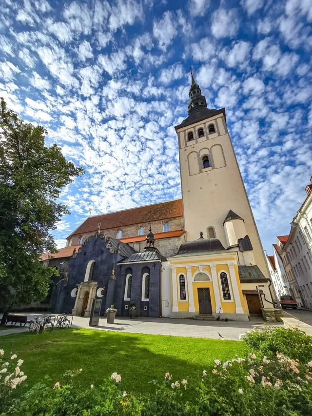 A pale church tower rises above a lawn, flower beds and a yellow doorway.