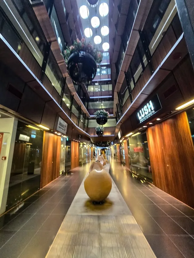 Hanging globe lights illuminate a covered shopping passage with smooth sculptures.
