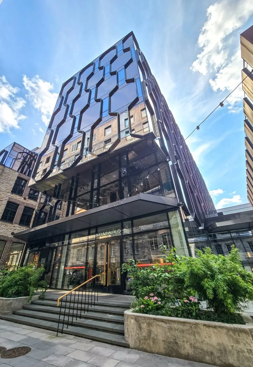 Angular reflective glass panels cover a modern building above steps and planted concrete containers.