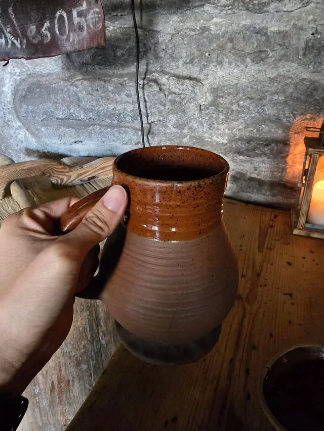 A hand holds an earthenware cup beside a lantern and a stone wall.