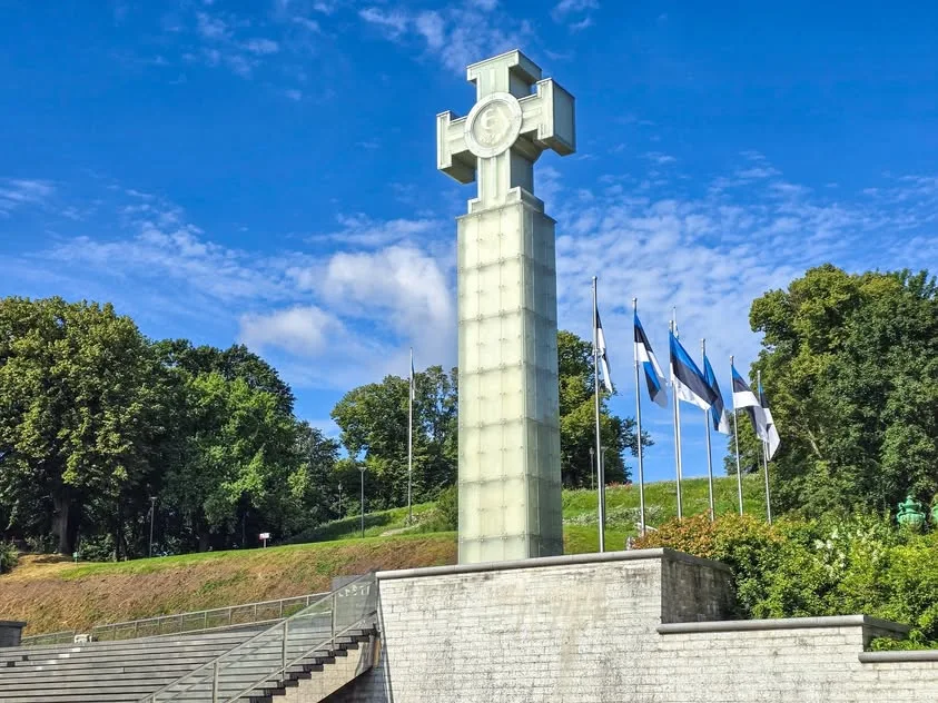 A translucent cross stands above broad steps beside Estonian flags.