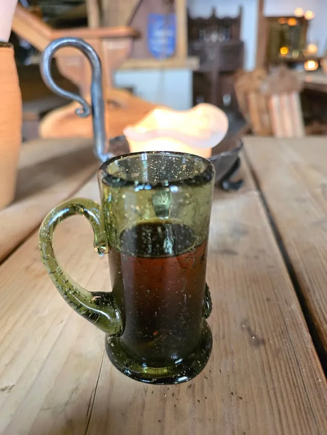 A green handled glass holds an amber drink beside a candle on a wooden table.