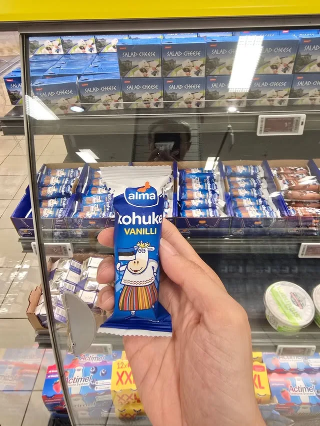 A blue Alma kohuke packet is held in front of a refrigerated dairy display.