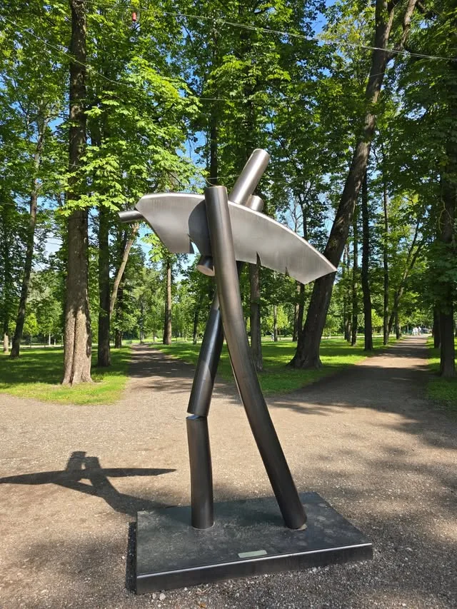 A geometric metal sculpture rises from a paved path beneath trees.