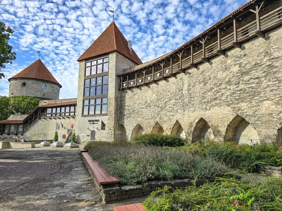 Stone towers with red tiled roofs connect to a high wall and a covered timber walkway above planted beds.