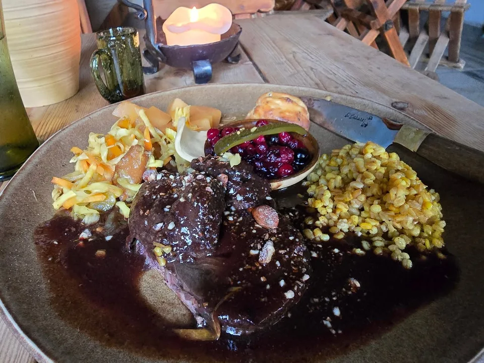 A main course in dark sauce sits beside grains, vegetables and berries on a wooden table with a candle.