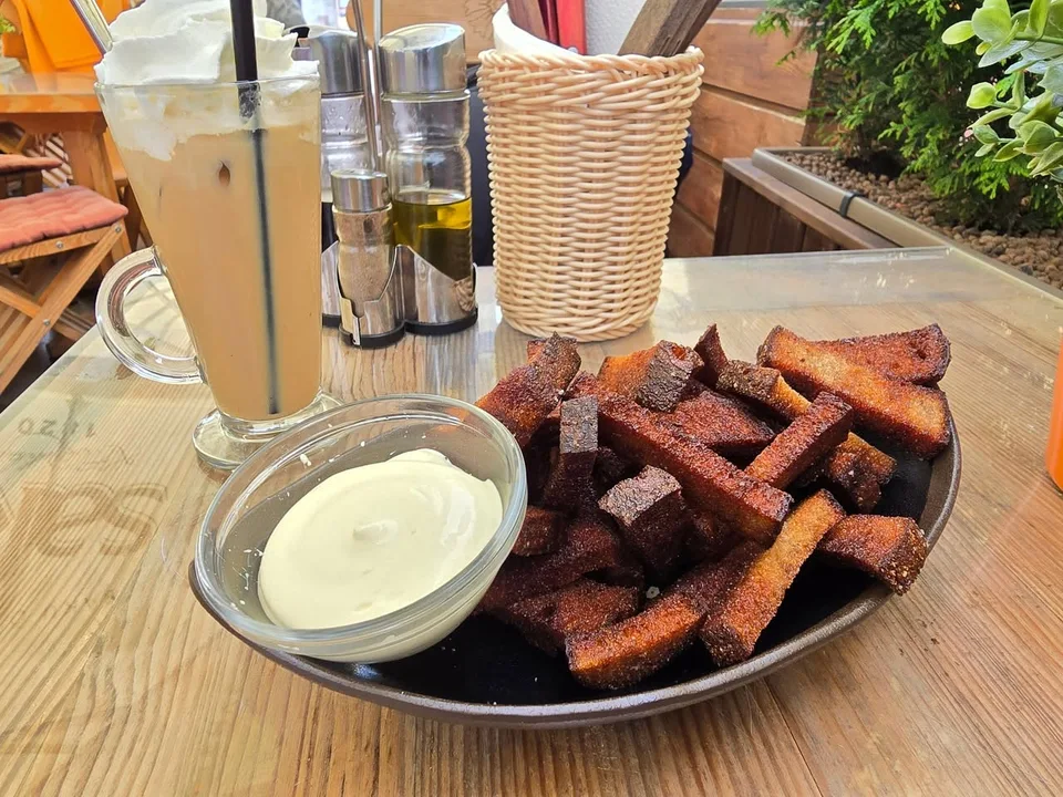 Browned strips of bread fill a plate beside a pale dip and a tall iced drink on a terrace table.