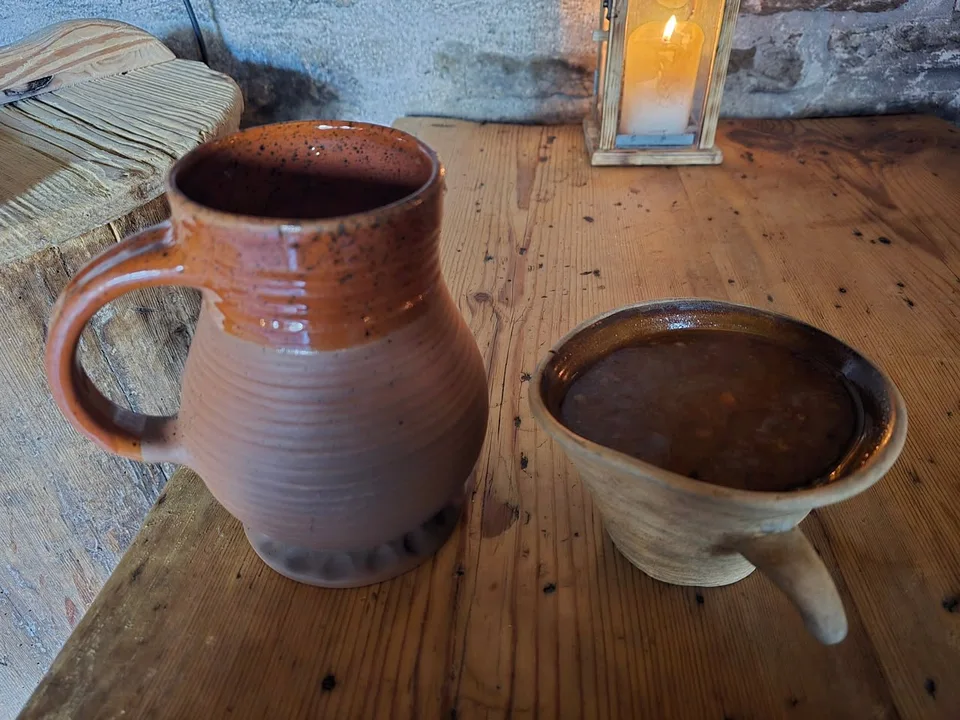 A brown earthenware jug and a handled bowl of soup stand on a wooden table beside a candle lantern.