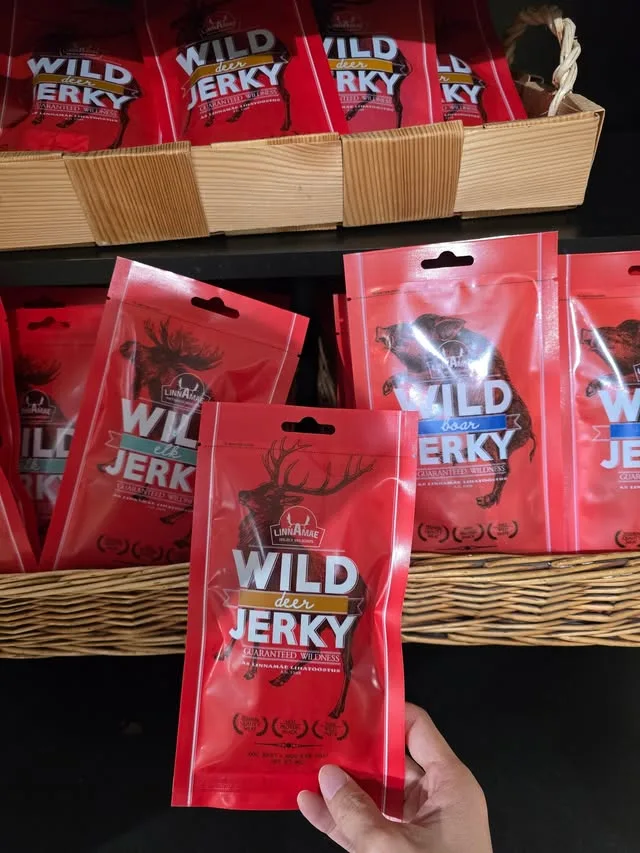 A packet of wild deer jerky is held in front of a shelf of similar packets.