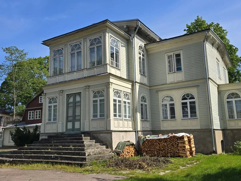 A pale wooden house with tall windows stands beside stacks of firewood.