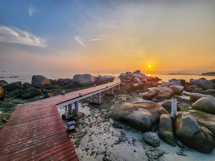 A wooden walkway with small lamps winds between coastal boulders towards the rising sun.