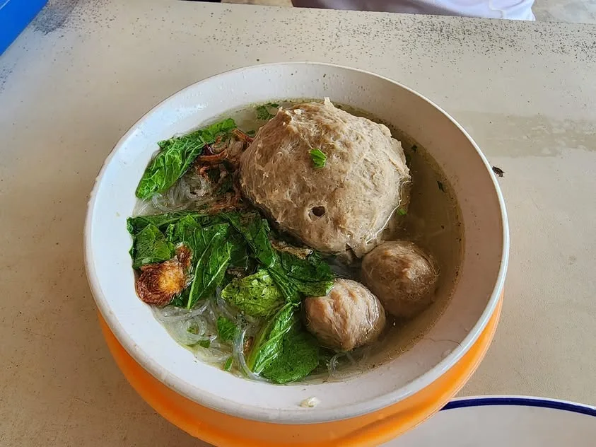 Meatballs, greens and fine noodles sit in a clear soup in a white bowl.