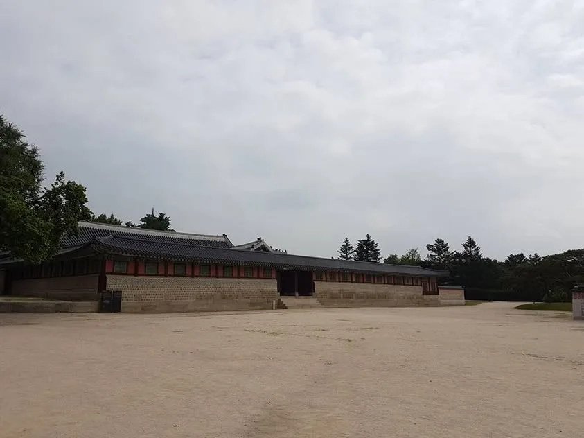 An empty sandy courtyard lies beside a low palace building with grey roof tiles and coloured shutters.