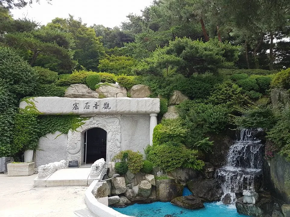A stone grotto entrance carved with dragons stands beside a small waterfall among green shrubs and pines.