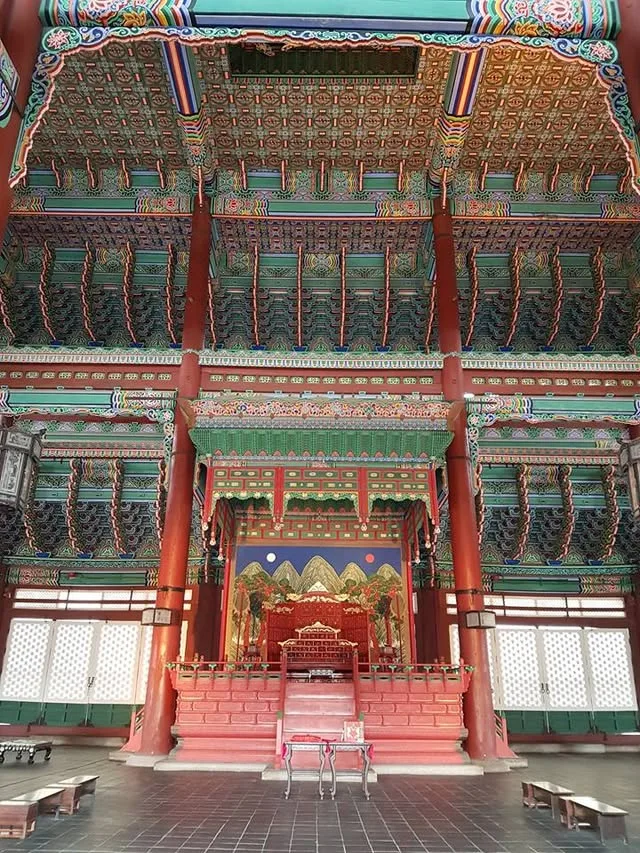 A red throne stands below painted beams, with a screen showing the sun, moon and five mountain peaks behind it.