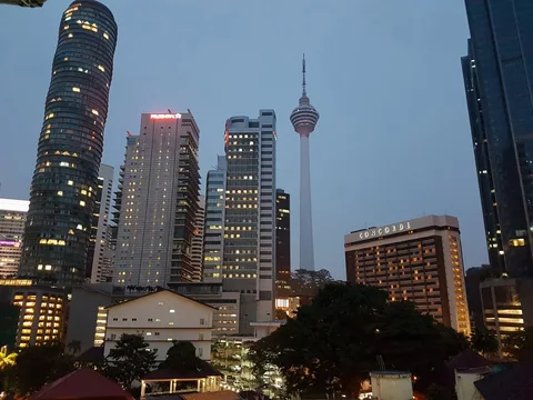 Kuala Lumpur Tower rises among illuminated buildings beneath a blue evening sky.