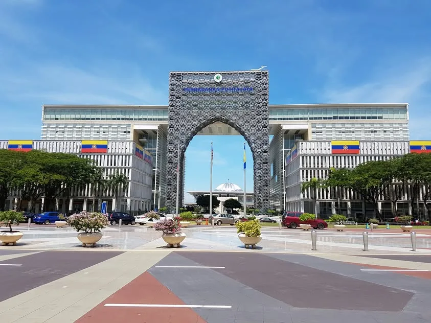 A tall patterned gateway bearing the Perbadanan Putrajaya name stands between long building wings, beyond cars and flower planters.