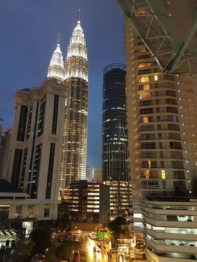 The illuminated Petronas Twin Towers rise behind nearby buildings and a curving road at night.