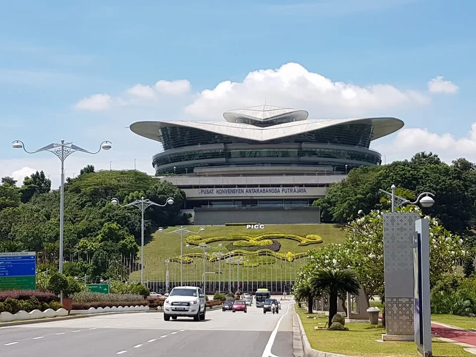 The broad curved roof of Putrajaya International Convention Centre sits above a landscaped hill and approach road.