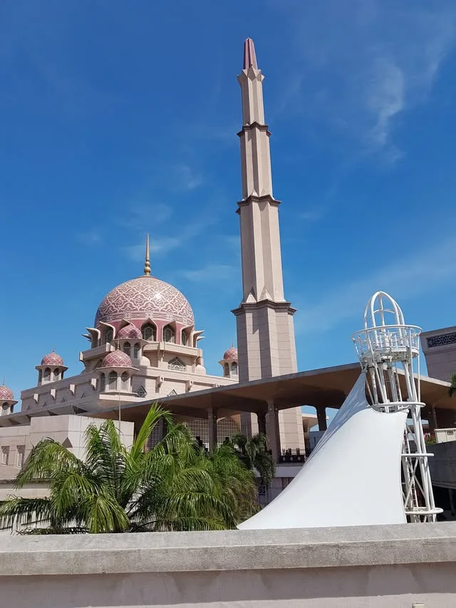 Putra Mosque’s pink patterned dome and tall minaret rise above palms and a covered walkway.