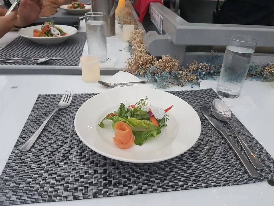 A white plate of salad sits on a woven placemat beside a water glass, small candle and dried flowers.