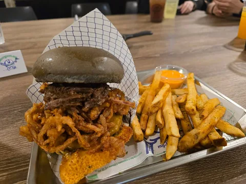 A tall burger in a dark bun sits beside fries and an orange dip on a metal tray.
