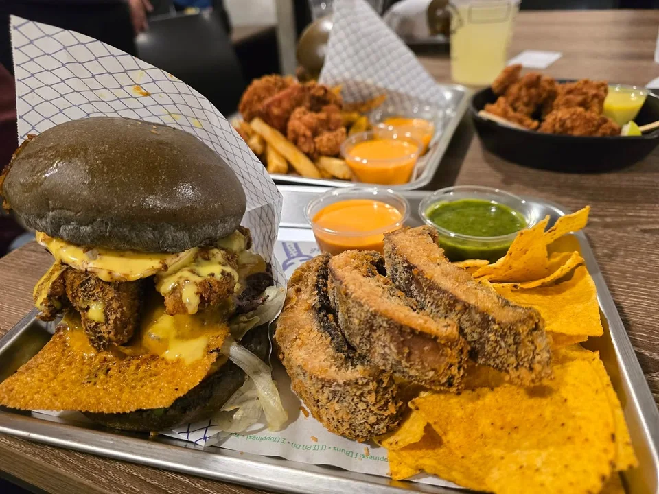 A dark-bun burger shares a tray with fried sides, yellow chips and orange and green dips.