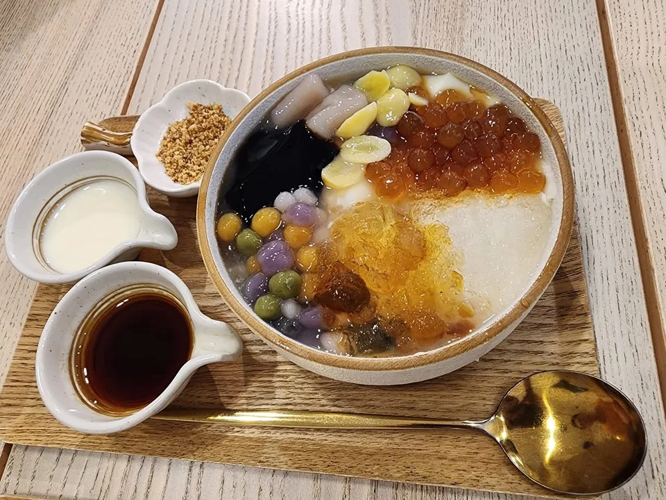 Shaved ice, dark jelly and colourful round toppings fill a bowl beside small jugs and a golden spoon.