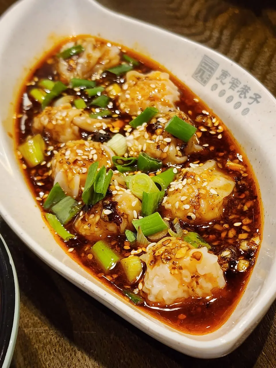 Dumplings sit in red sauce topped with sesame seeds and chopped spring onions in a white oval bowl.