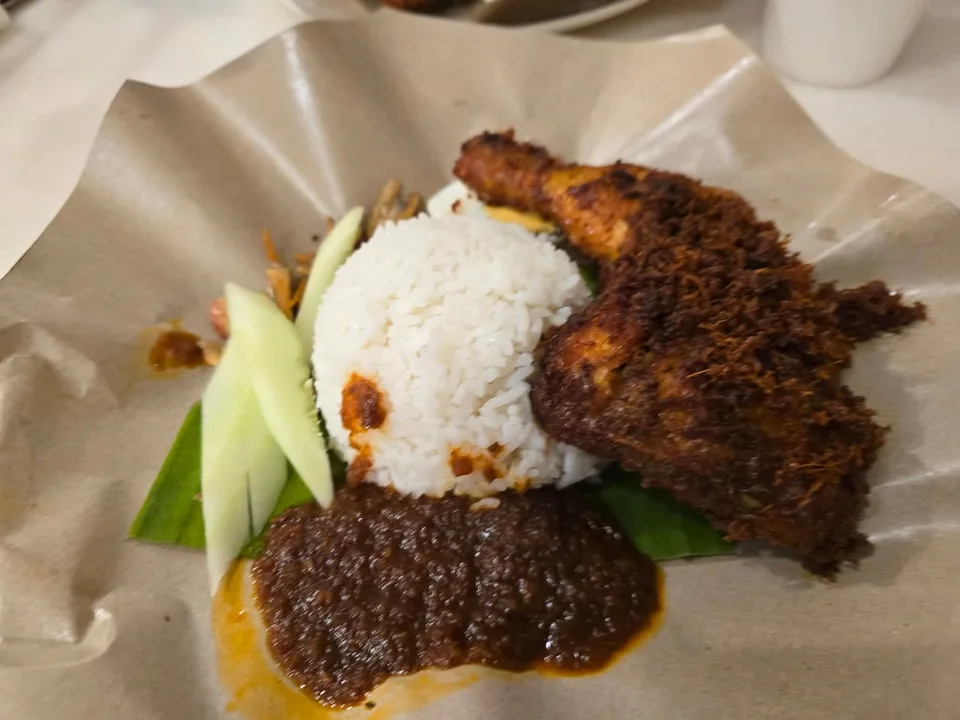 A mound of white rice sits beside a browned chicken leg, cucumber and dark sauce on a paper-lined plate.