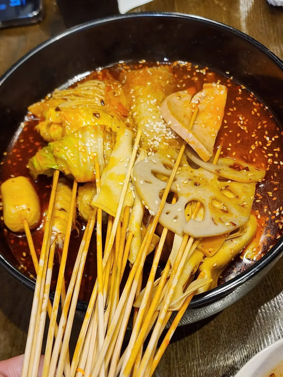 Wooden skewers hold lotus-root slices and folded vegetables in a black bowl of red broth.