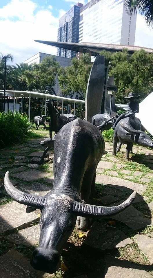 Dark carabao sculptures stand on a grassy patch beside a pale railing, with a sculpted farmer sitting on one animal.