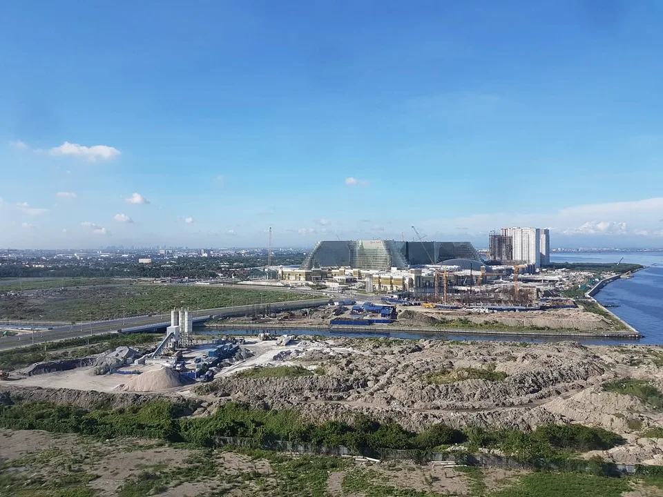 An elevated view across construction sites and roads beside Manila Bay, with a large resort complex in the distance.