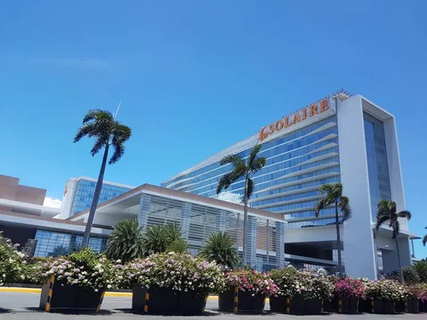 The Solaire building rises behind palm trees and large flower planters beneath a clear blue sky.