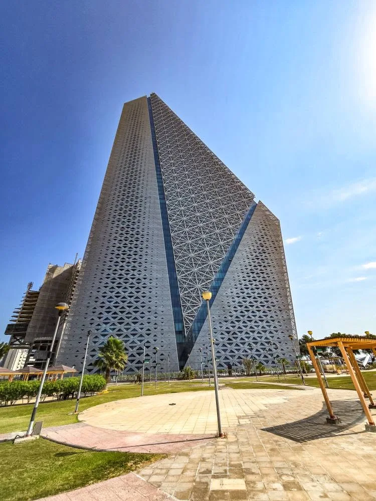 A tall tower covered in repeating triangular patterns rises above trees and a lawn.